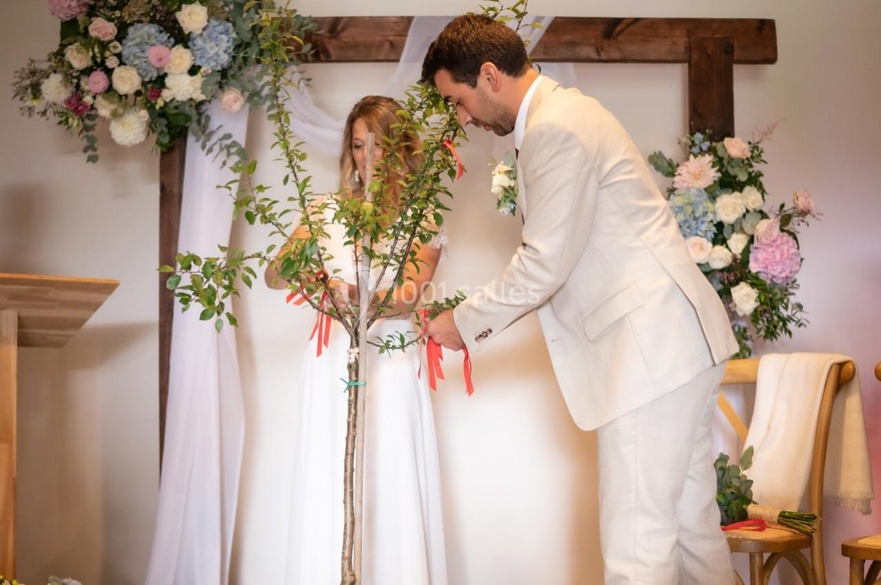 Un couple attache des rubans rouges à un jeune arbre lors d'une cérémonie, entouré de fleurs et de décorations blanches.