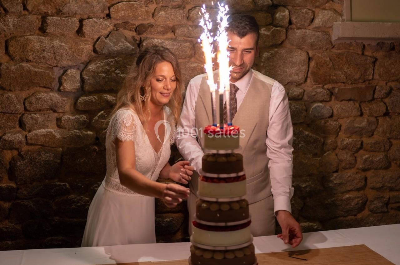 Un couple coupe un gâteau de mariage décoré de bougies étincelantes, devant un mur en pierre.