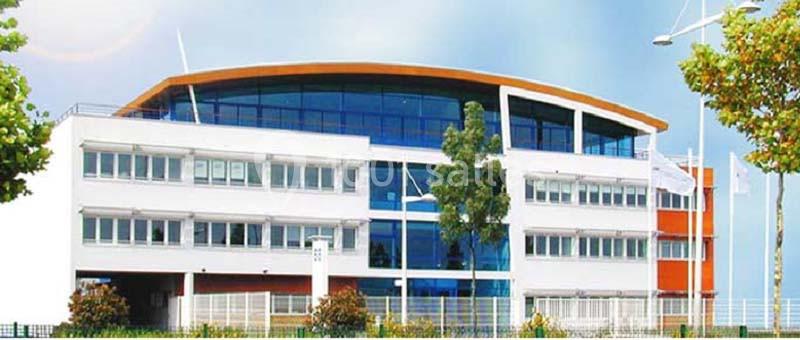 Bâtiment moderne à trois étages avec façade vitrée, entouré d'arbres et d'un ciel dégagé.