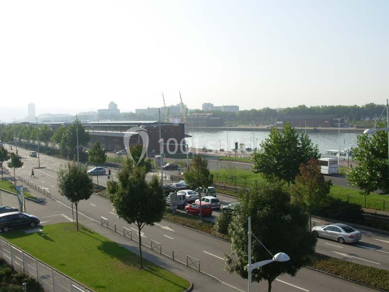 Vue d'une route bordée d'arbres avec un parking, un fleuve et des bâtiments en arrière-plan par temps clair.