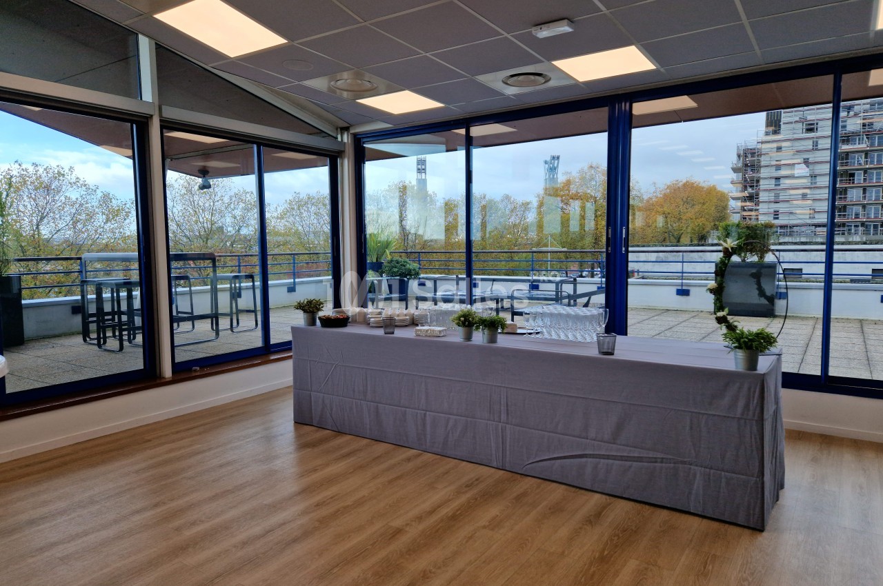 Salle lumineuse avec une table recouverte d'une nappe grise, vue sur une terrasse et des arbres à travers de grandes baies…