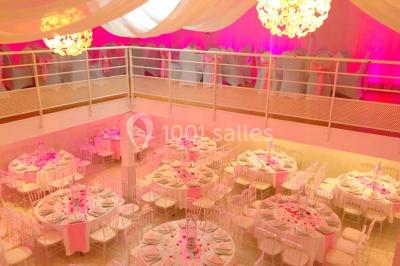 Salle de réception décorée avec des tables rondes, nappes blanches, chaises transparentes et centres de table fleuris.
