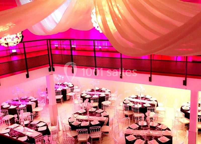 Salle de réception décorée avec des tables rondes, nappes noires et blanches, chaises transparentes et drapés au plafond.