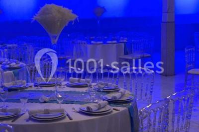 Salle de réception décorée avec des tables rondes, nappes blanches, chaises transparentes et centres de table fleuris.