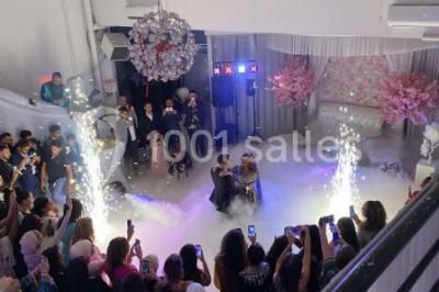 Salle de réception décorée avec des tables rondes, nappes blanches, chaises transparentes et centres de table fleuris.