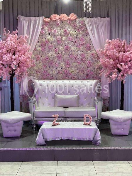 Décor floral avec canapé blanc capitonné, deux tabourets, rideaux roses et arbres ornés de fleurs roses.