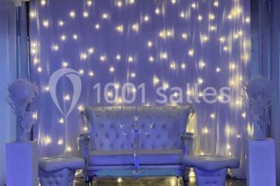 Salle de réception décorée avec des tables rondes, nappes blanches, chaises transparentes et centres de table fleuris.