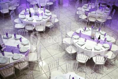 Salle de réception décorée avec des tables rondes, nappes blanches, chaises transparentes et centres de table fleuris.