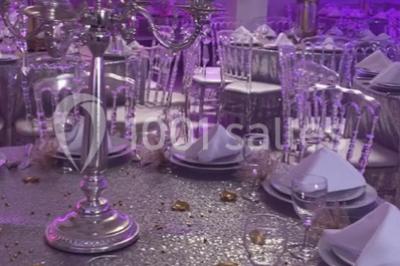Salle de réception décorée avec des tables rondes, nappes blanches, chaises transparentes et centres de table fleuris.