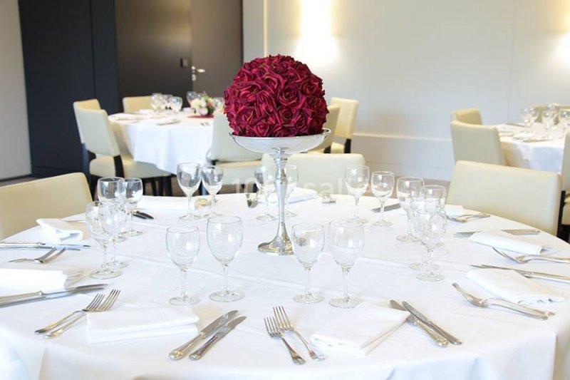 Table ronde élégamment dressée avec nappes blanches, vaisselle raffinée et centre de table floral rouge.