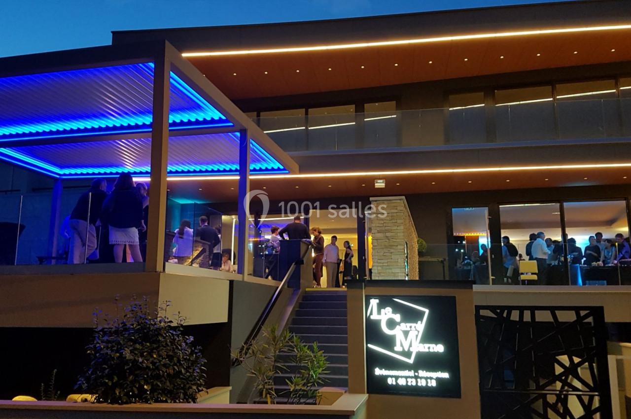 Façade illuminée d'un restaurant moderne en soirée, avec des invités visibles à l'intérieur et sur la terrasse.