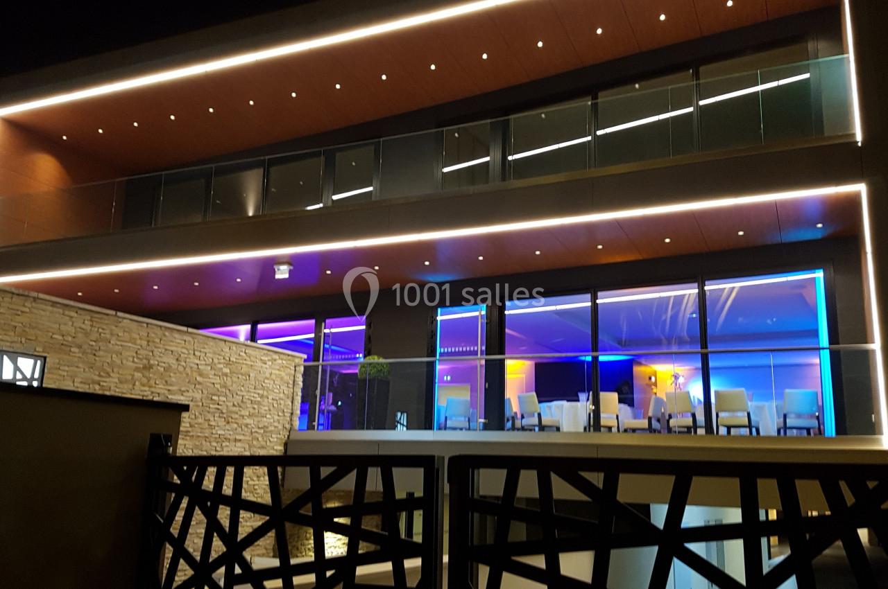 Façade moderne d'un bâtiment éclairé par des lumières colorées, avec une terrasse vitrée et un mur en pierre.