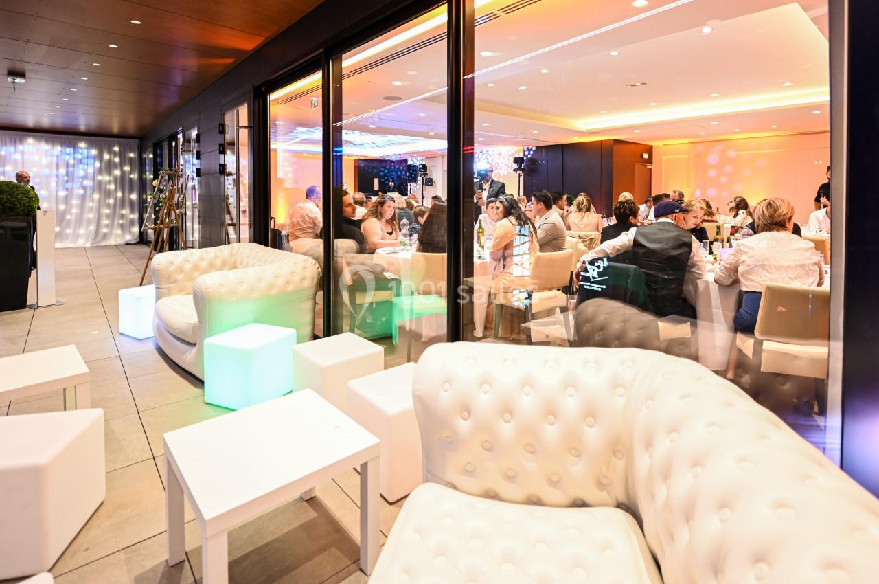 Salle de réception avec des invités assis à l'intérieur, vue depuis une terrasse avec fauteuils et tables lumineuses.