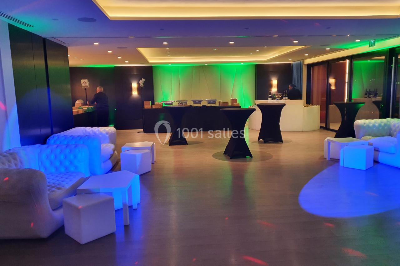 Salle de réception moderne avec éclairage coloré, tables hautes noires et canapés blancs disposés autour.