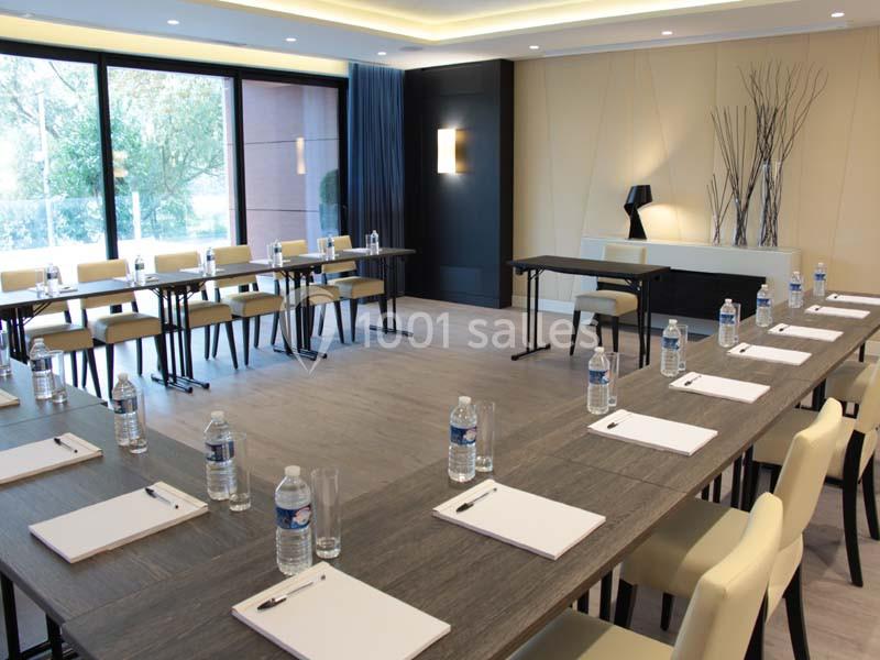 Salle de réunion moderne avec tables disposées en U, chaises, blocs-notes, bouteilles d'eau et grande baie vitrée.