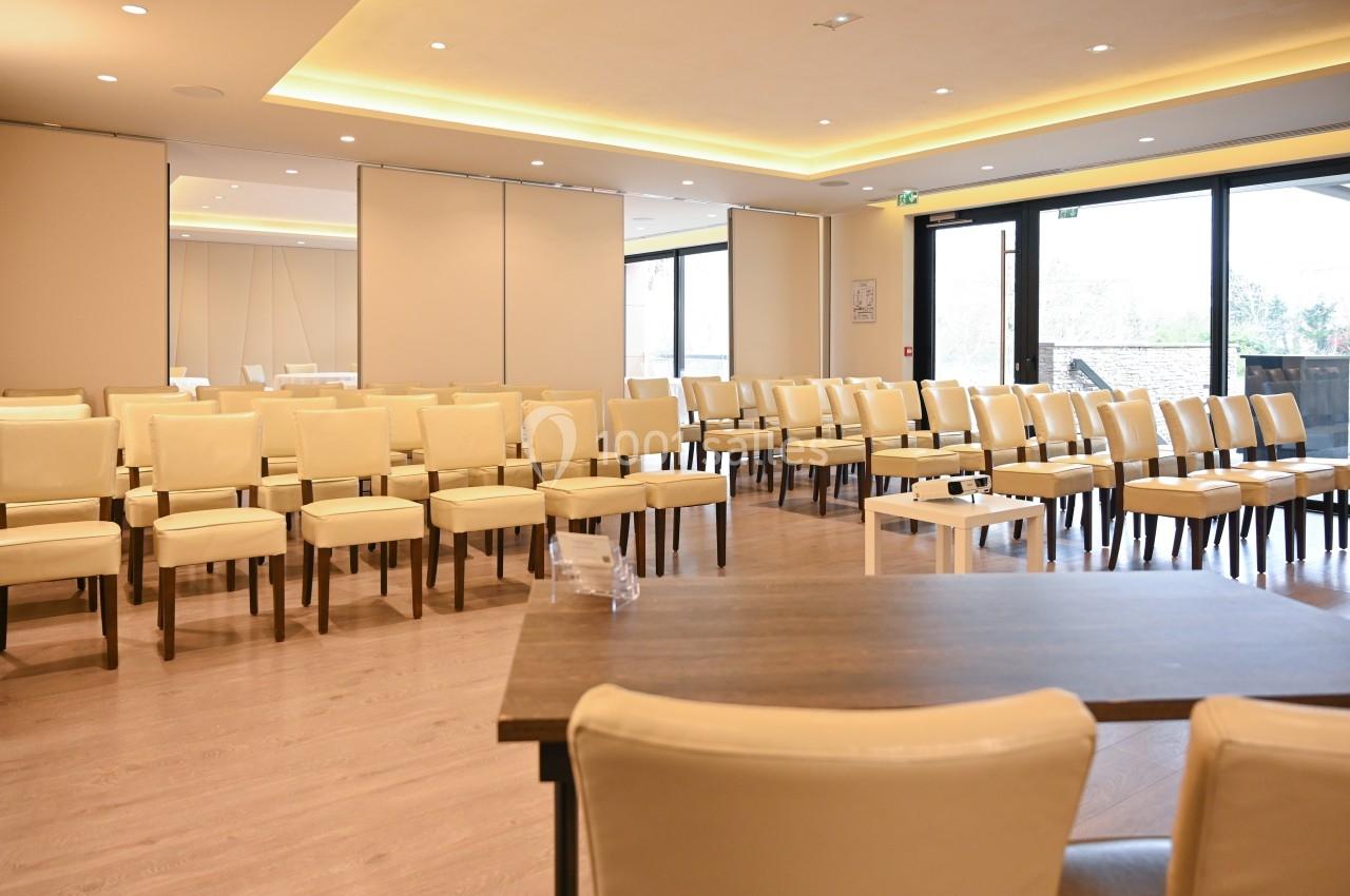 Salle de conférence lumineuse avec des chaises beige alignées, tables en bois et grandes baies vitrées donnant sur l…