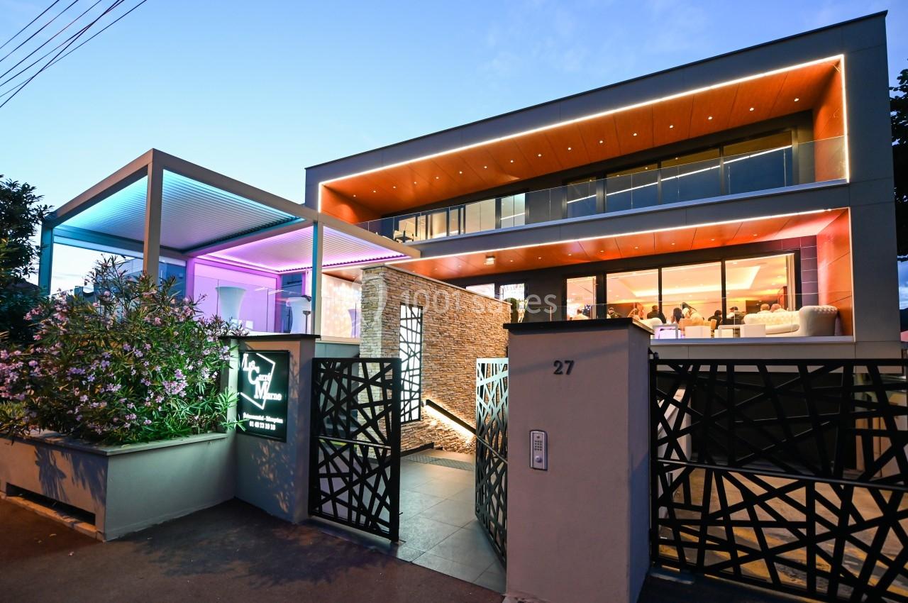 Façade moderne d'un bâtiment éclairé par des lumières colorées au crépuscule, avec une entrée design et végétation.