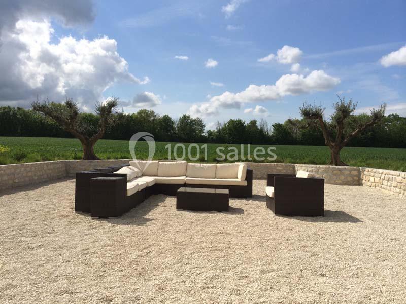 Location salle Benon (Charente-Maritime) - Abbaye de la Grâce-Dieu #8 Salon de jardin en résine tressée avec coussins beiges, installé sur une terrasse en gravier, entourée de verdure.