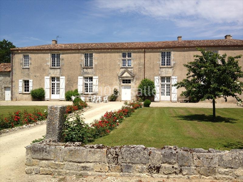 Location salle Benon (Charente-Maritime) - Abbaye de la Grâce-Dieu #1 Façade d'une grande maison en pierre avec volets blancs, jardin fleuri et allée gravillonnée sous un ciel dégagé.