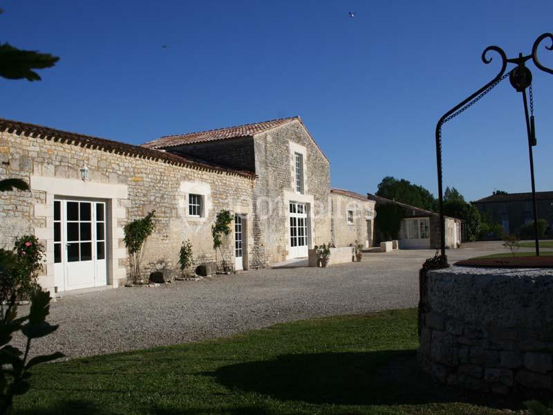 Location salle Benon (Charente-Maritime) - Abbaye de la Grâce-Dieu #3 Bâtiment en pierre avec des portes vitrées, entouré de végétation et d'une cour gravillonnée sous un ciel bleu.