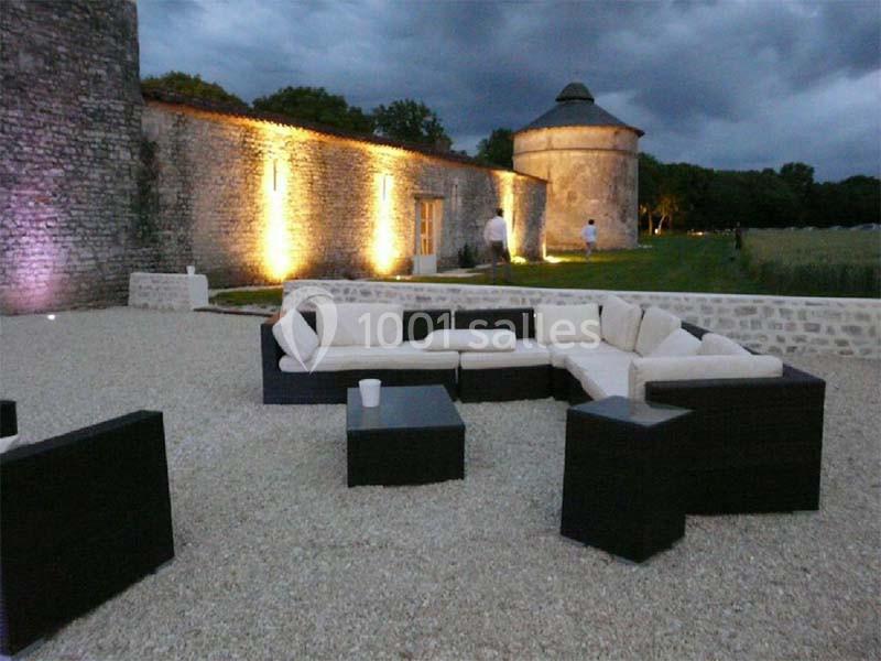 Location salle Benon (Charente-Maritime) - Abbaye de la Grâce-Dieu #6 Mobilier de jardin moderne avec canapés et tables basses sur une terrasse en gravier, près d'un mur en pierre éclairé.