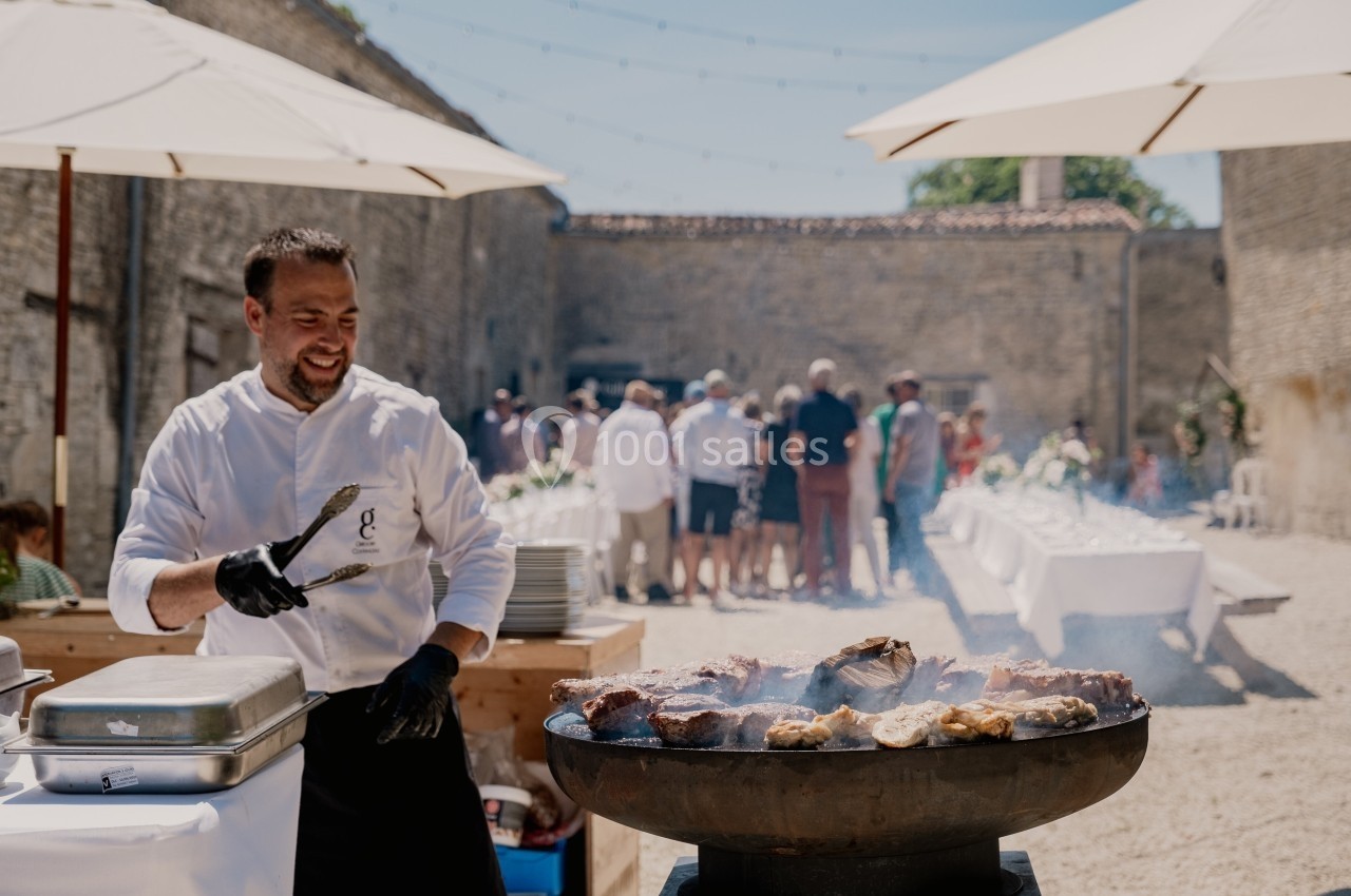 Location salle Benon (Charente-Maritime) - Abbaye de la Grâce-Dieu #11 Un chef cuisine des viandes sur un barbecue en plein air lors d'un événement avec des invités en arrière-plan.