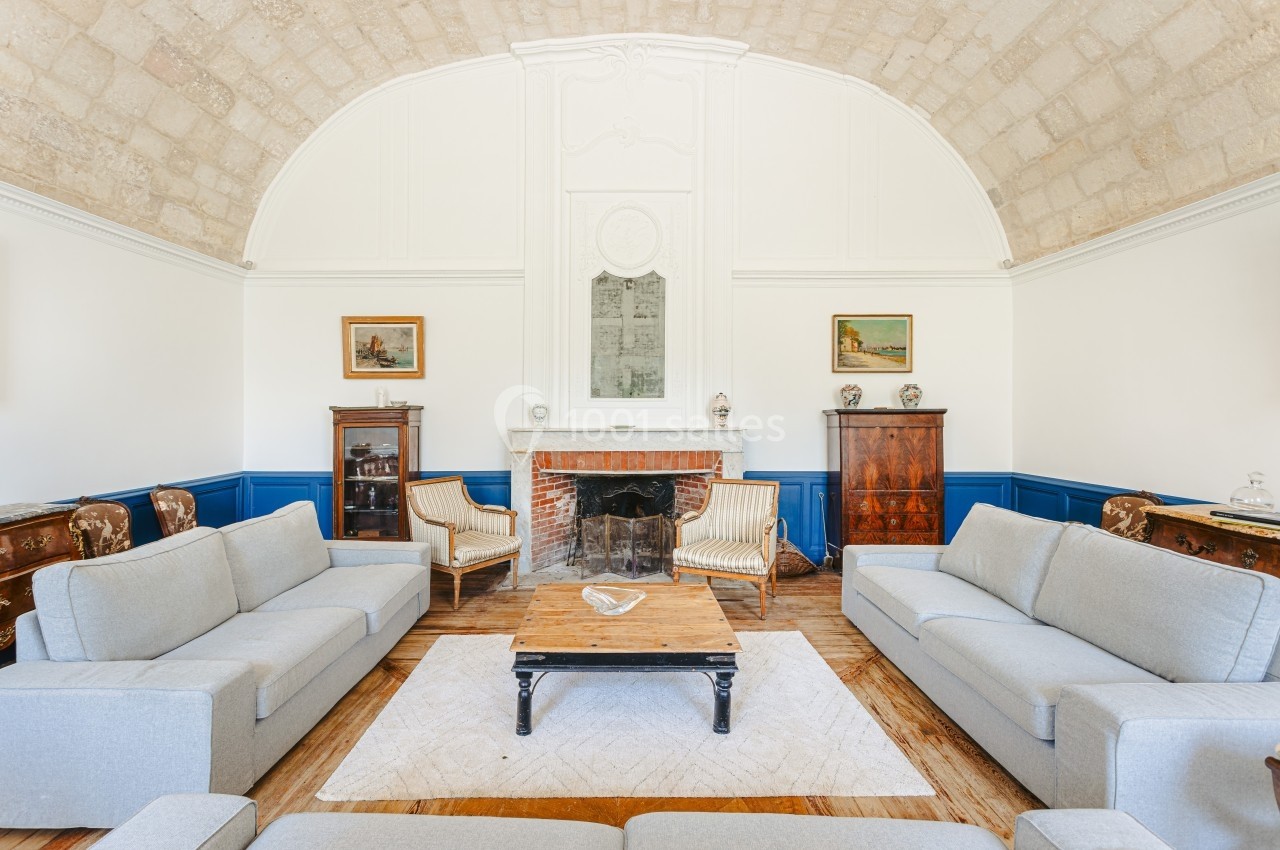 Location salle Benon (Charente-Maritime) - Abbaye de la Grâce-Dieu #19 Salon lumineux avec murs blancs, plafond voûté, cheminée centrale, meubles anciens et canapés gris.