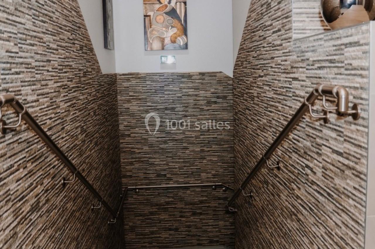 Escalier intérieur avec murs en pierre, mains courantes métalliques et tableau décoratif au mur du fond.