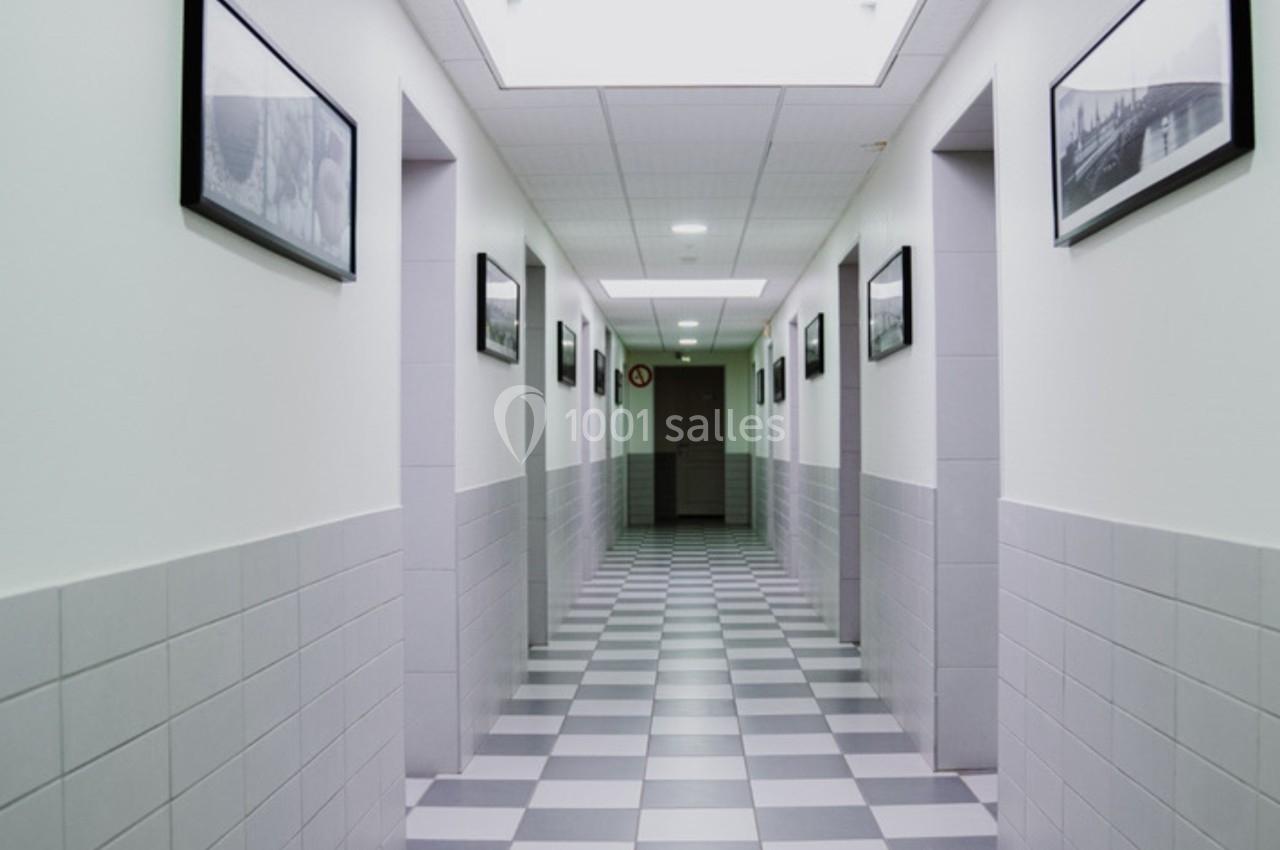 Couloir intérieur avec carrelage à motifs en damier, murs blancs ornés de cadres noirs et éclairage au plafond.