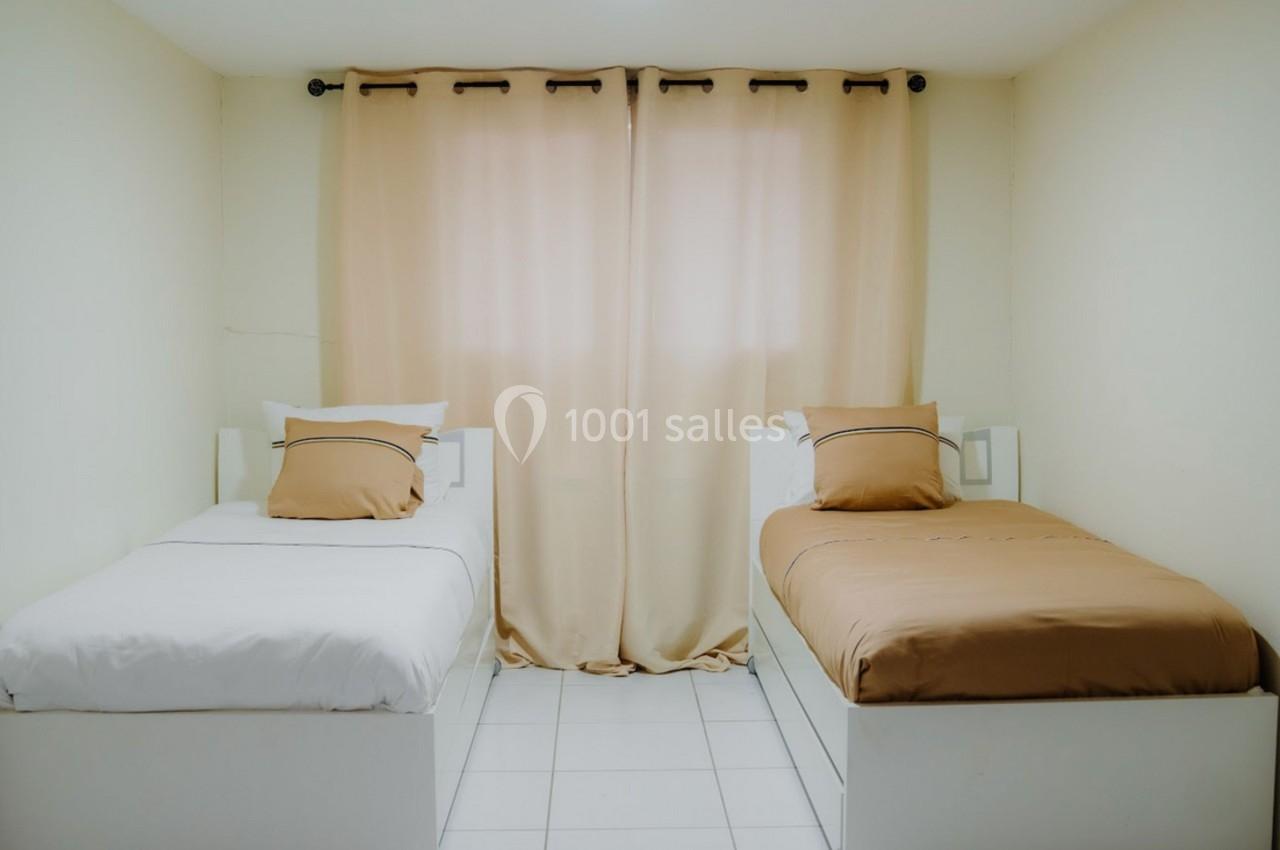 Deux lits simples avec draps blancs et beiges dans une chambre lumineuse aux murs clairs et rideaux beige clair.