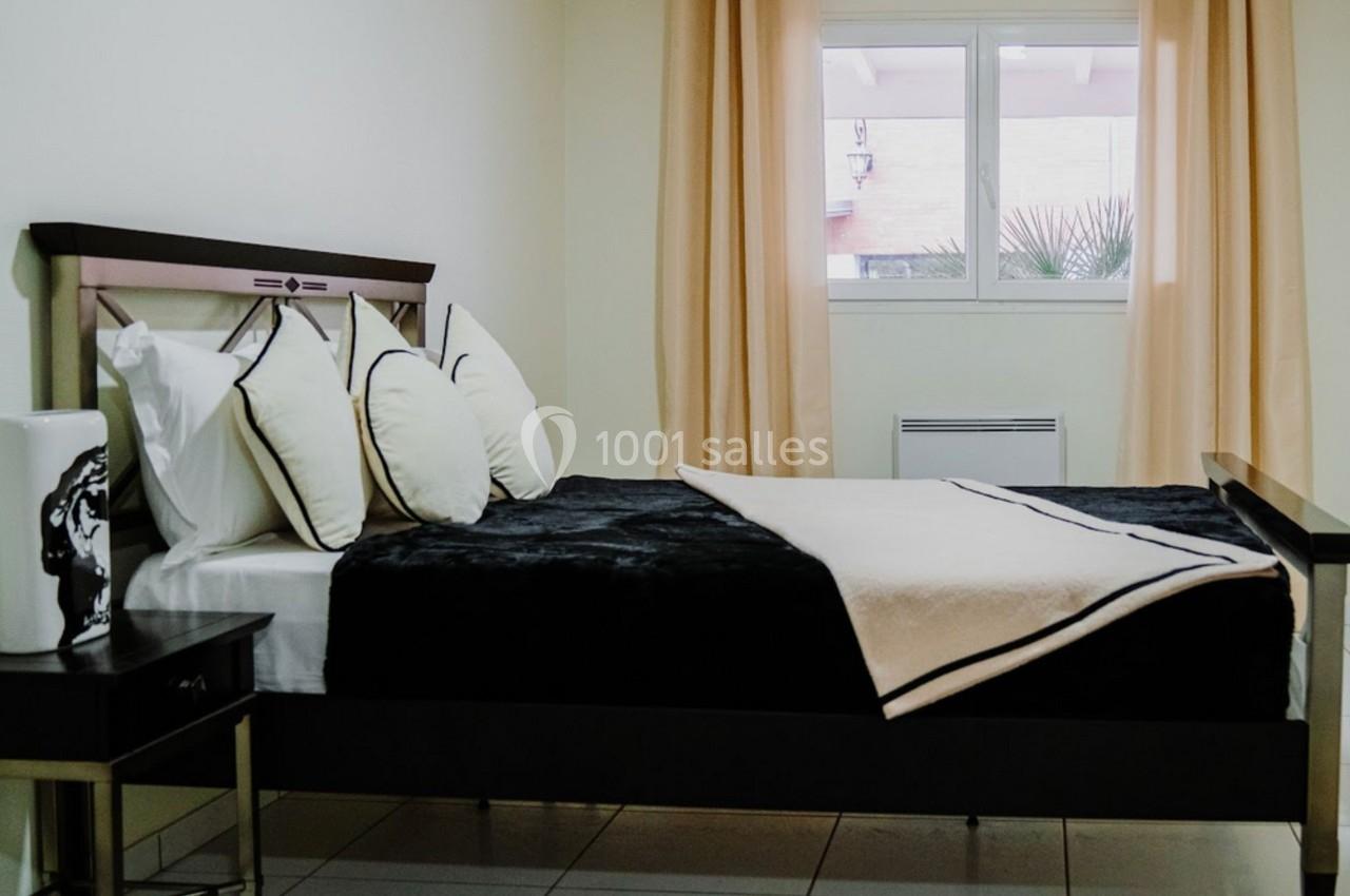 Chambre lumineuse avec un lit simple, draps noirs et beige, tapis zébré et fenêtre encadrée de rideaux beige clair.