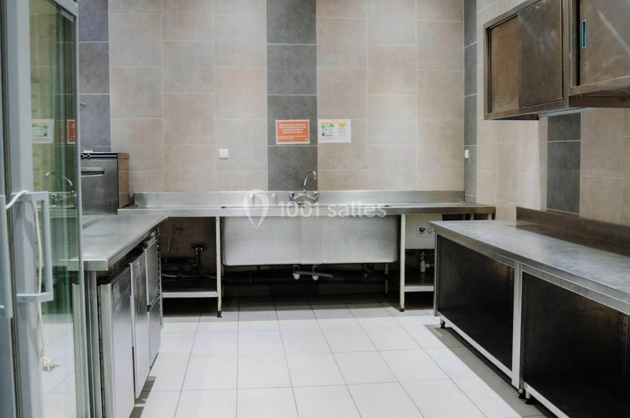 Cuisine professionnelle avec plans de travail en inox, évier central, réfrigérateur vitré et murs carrelés.