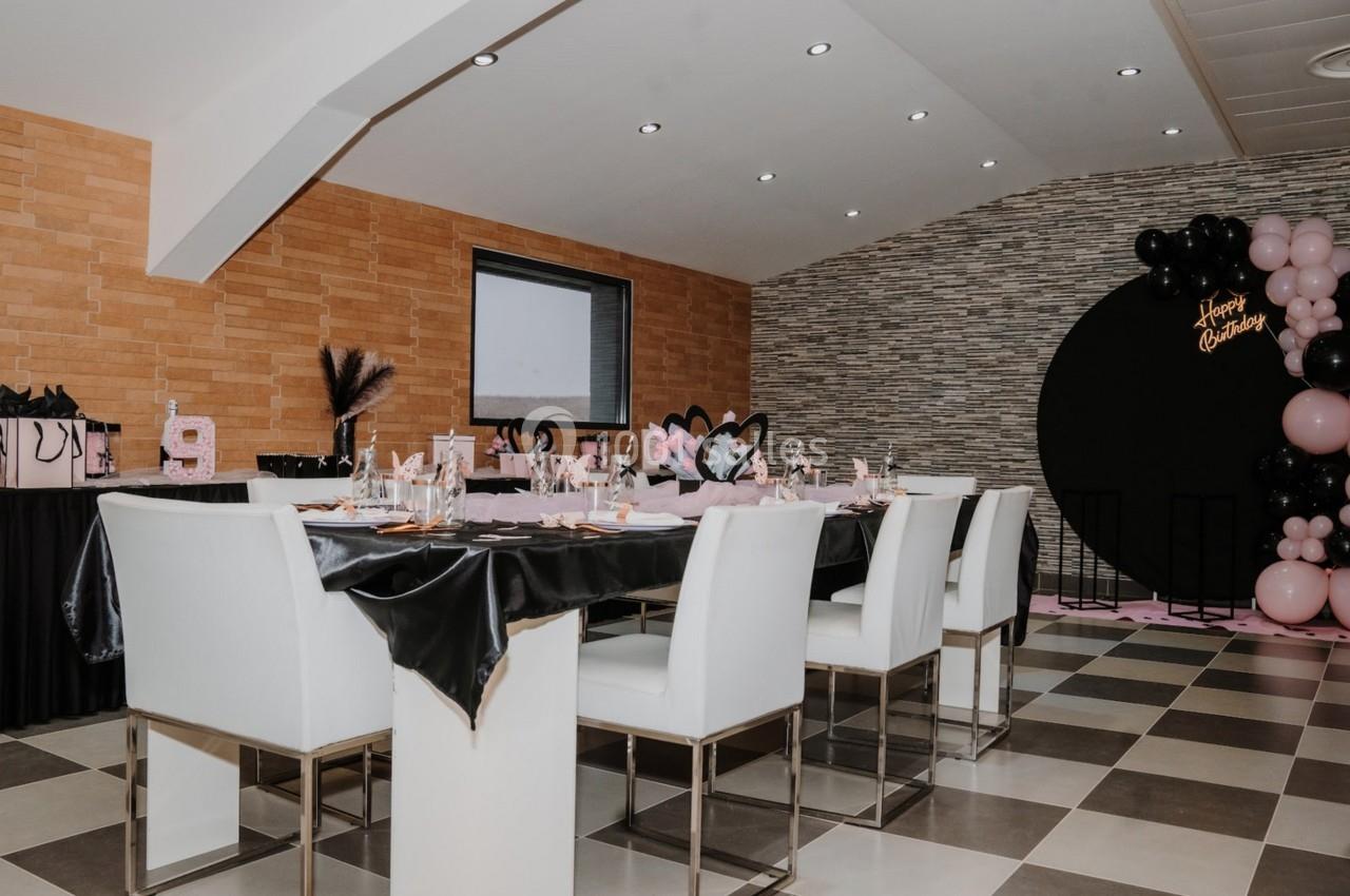 Salle décorée pour une fête avec table dressée, ballons roses et noirs, et cadeaux sur une table latérale.