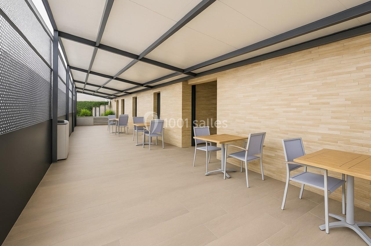 Terrasse couverte avec tables et chaises en métal gris, murs en briques claires et sol en carrelage beige.