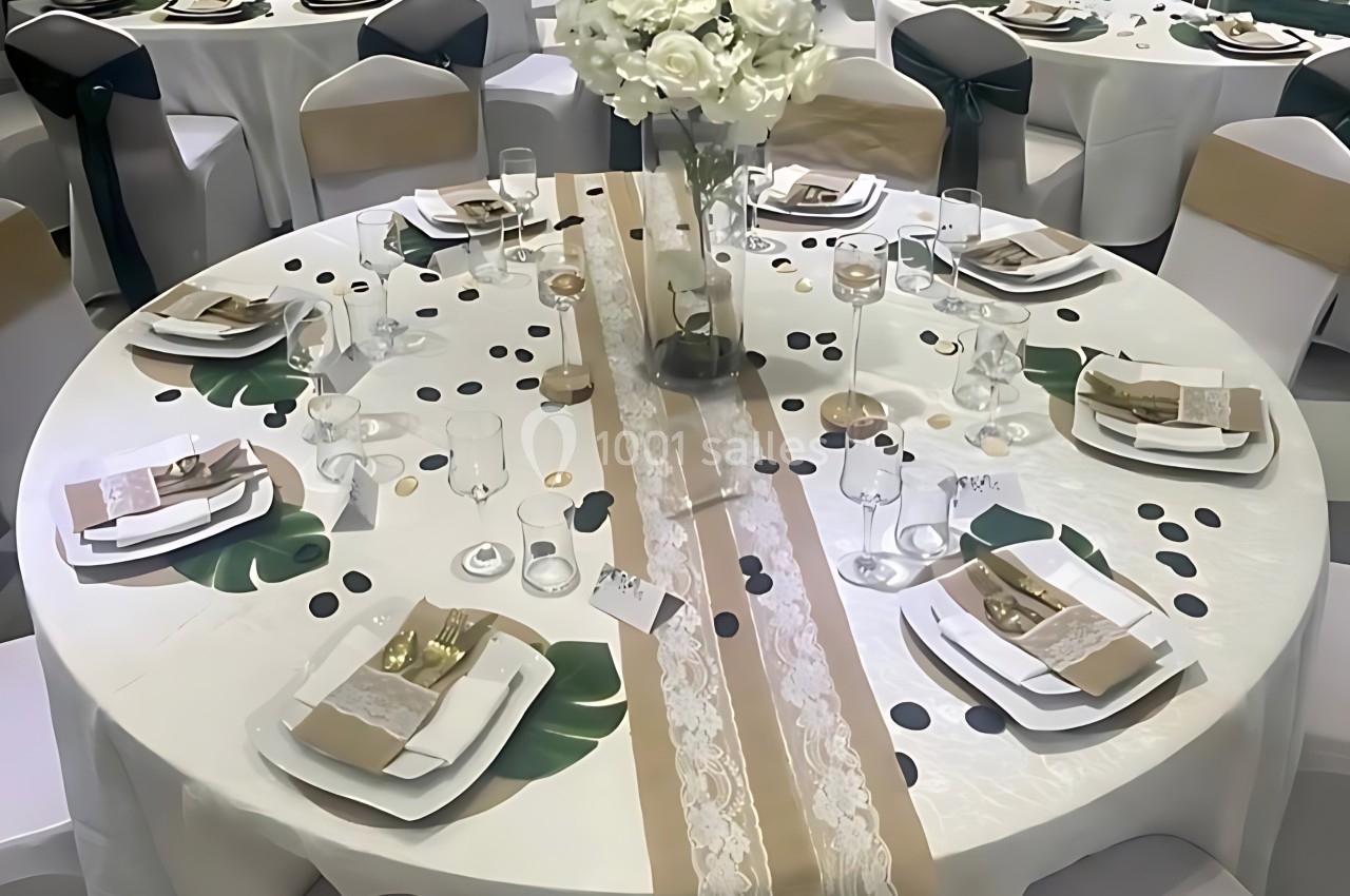 Table ronde décorée pour un événement, avec nappe blanche, chemins de table beige et blanc, et centre de table floral.