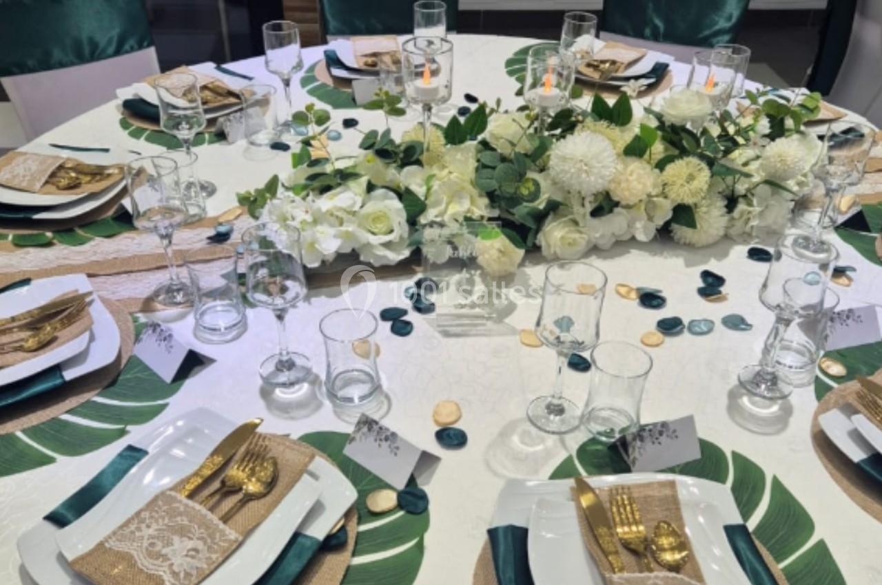 Table ronde élégamment dressée avec vaisselle blanche, couverts dorés, décorations florales blanches et feuillages verts.