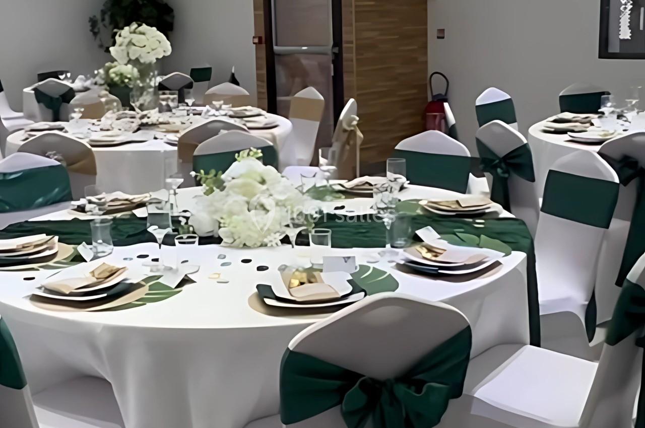 Salle décorée pour un événement, avec tables rondes dressées, nappes blanches et chaises ornées de nœuds verts.