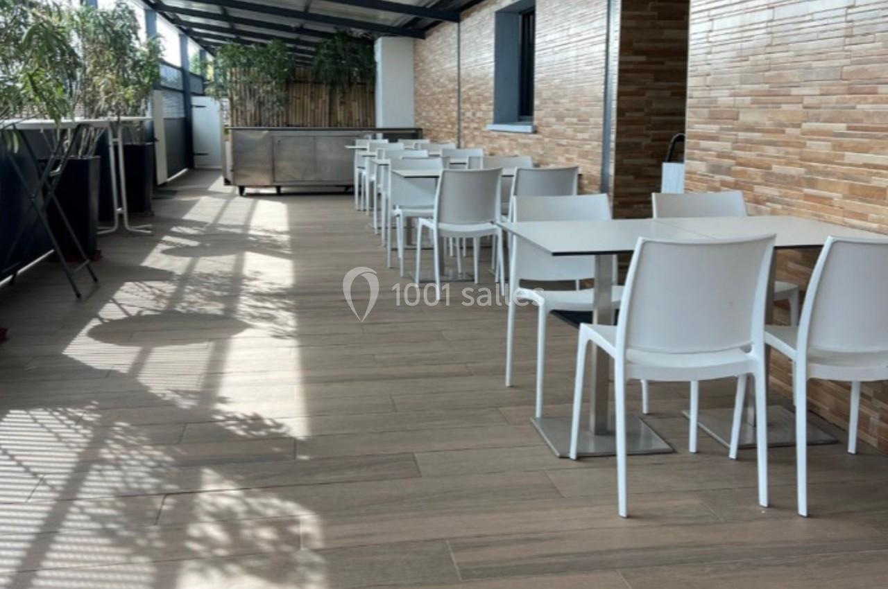 Terrasse couverte avec sol en bois, tables blanches alignées et murs en briques claires, éclairée par la lumière naturelle.