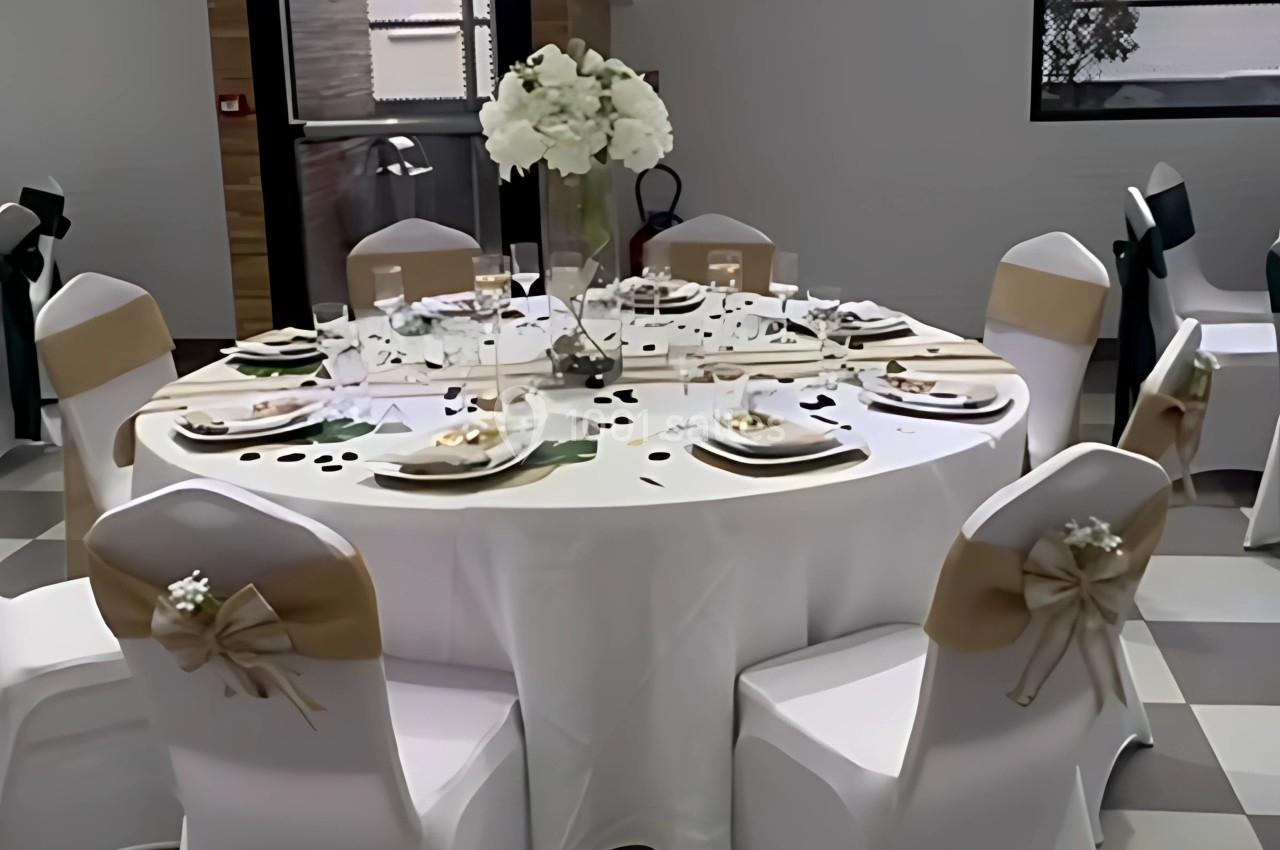Table ronde décorée pour un événement, avec nappes blanches, chaises ornées de nœuds beige et centre de table floral.