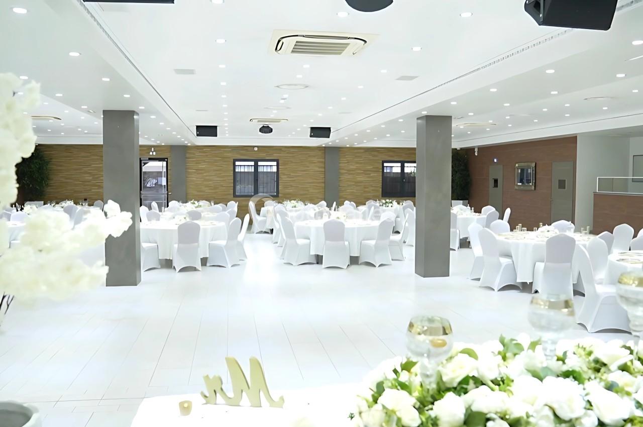 Salle de réception lumineuse avec des tables rondes dressées, des chaises blanches et des décorations florales.