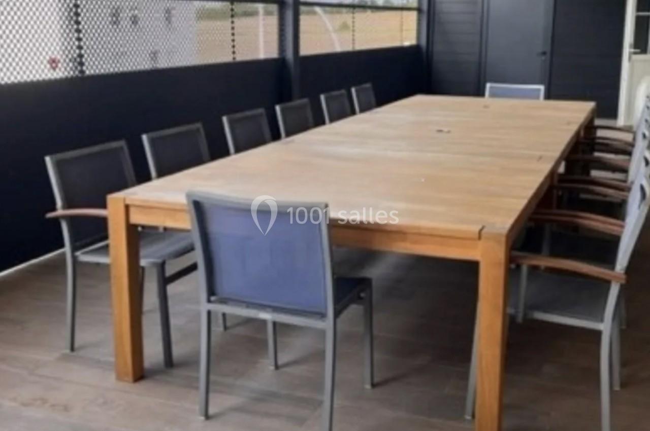 Table rectangulaire en bois entourée de chaises avec accoudoirs, située sur une terrasse couverte au sol carrelé.