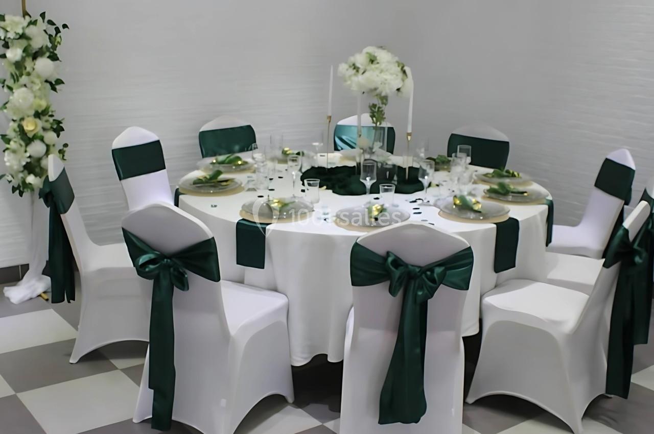Table ronde décorée pour un événement, nappes blanches, chaises avec nœuds verts et centre de table floral.