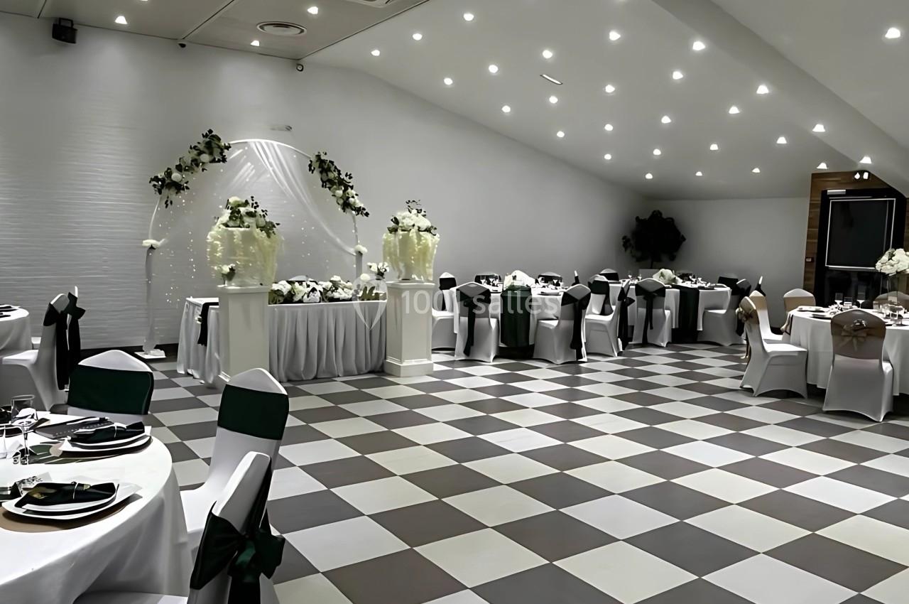 Salle de réception décorée pour un événement, avec tables dressées, chaises ornées et arche florale blanche.