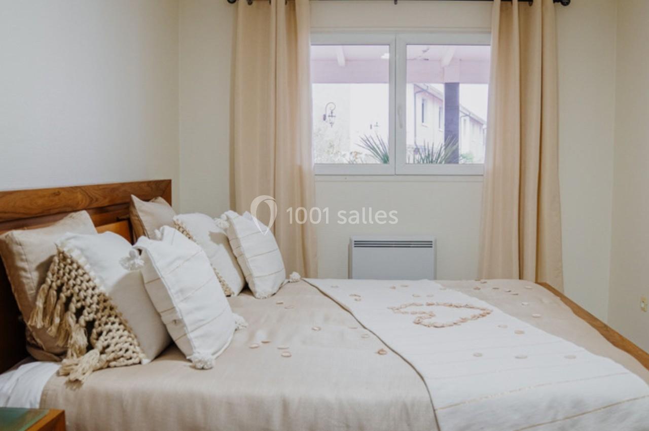 Chambre lumineuse avec lit double, draps beige clair, coussins décoratifs et fenêtre avec rideaux assortis.