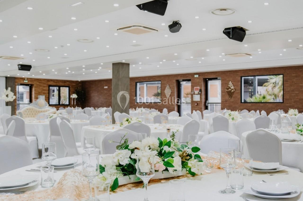 Salle de réception élégante avec tables rondes dressées, nappes blanches et décoration florale au centre.