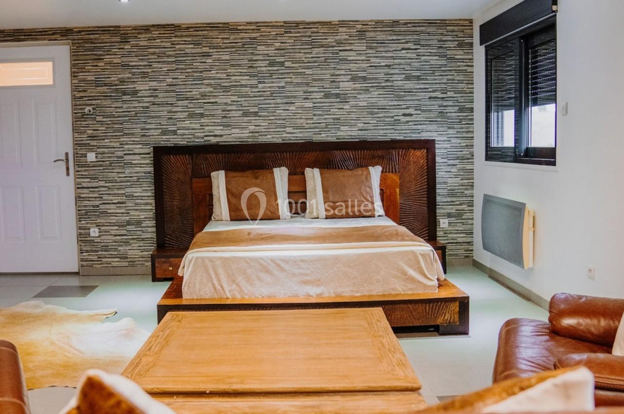 Chambre moderne avec lit double, mur en pierre, canapé en cuir et table basse en bois dans un espace lumineux.