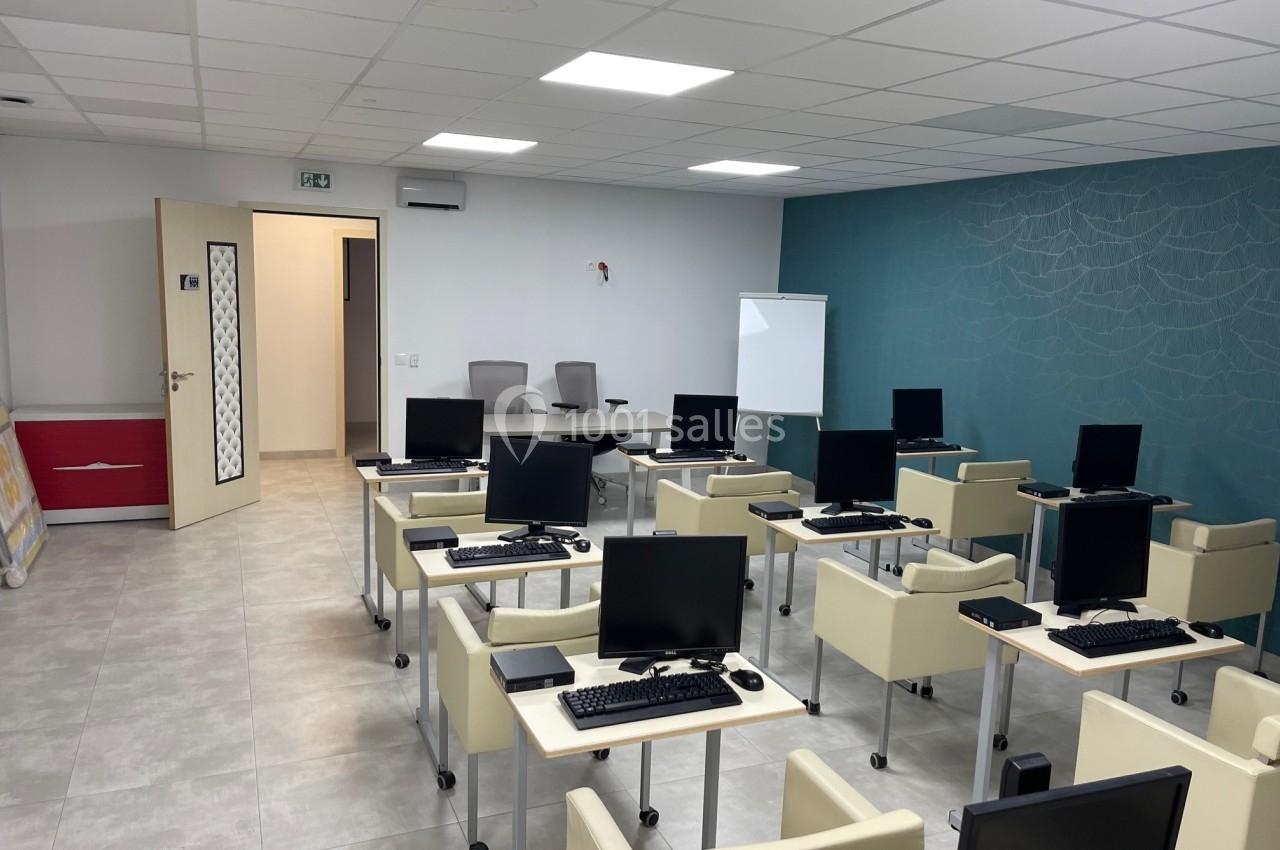 Salle de formation équipée de bureaux avec ordinateurs, chaises, tableau blanc et éclairage au plafond.