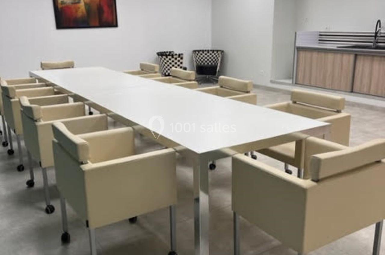 Salle de réunion avec une grande table blanche entourée de chaises beige sur roulettes, et un coin kitchenette en arrière…