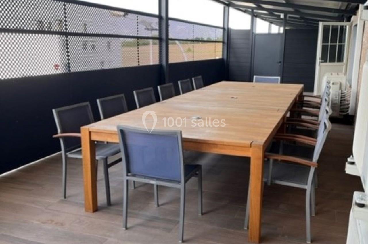 Table en bois rectangulaire entourée de chaises sur une terrasse couverte avec sol en bois et panneaux ajourés.