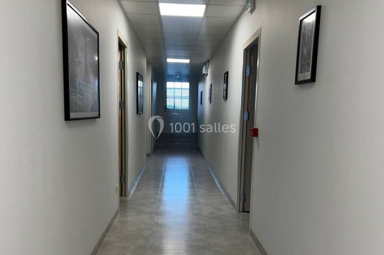 Couloir lumineux avec sol en carrelage, murs blancs ornés de cadres, et une fenêtre au fond.